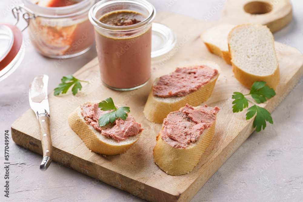 Homemade chicken liver pate on fresh french white wheat baguette slices on wooden plate, glass mason jars with cooked liverwurst, top view
