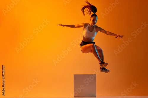 Muscular strong body. Young sportive girl, athlete training, jumping over block against orange studio background in neon light. Concept of sportive lifestyle, health, endurance, action and motion. Ad