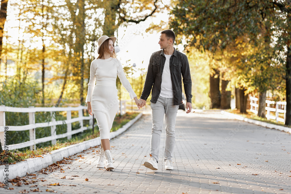 Fototapeta premium Pregnancy and Parenthoods concept - smiling man is walking with pregnant wife and looking to each other in autumn park. Happy styling young family is relaxing outdoors on weekend on sunny day.