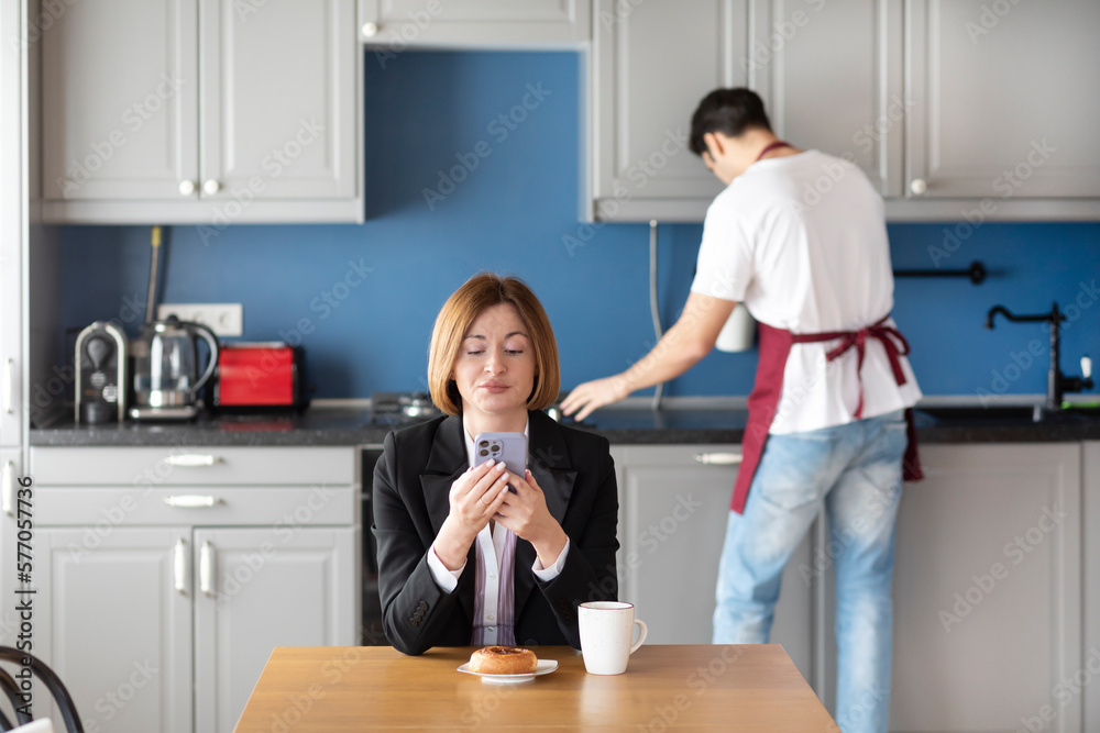 Confident business woman sits at the table and looking at her smartphone and her husband householder takes care about the house Confident business woman sits at the table and looking at her smartphone and her husband householder takes care about the house