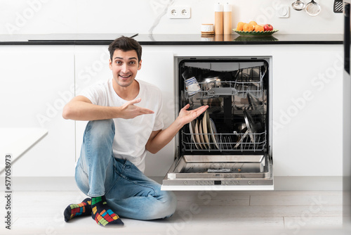 Young smiling man putting dirty plates in dishwasher machine in the kitchen. Household and exhausting cleaning day concept