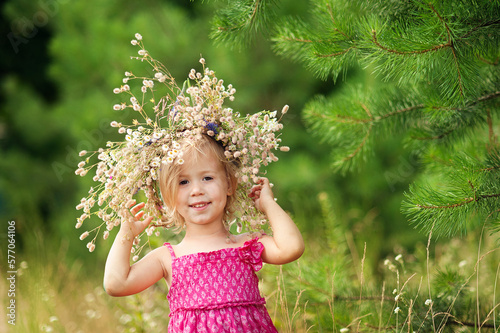little girl is wearing flower wreath on her head in field on summer sunny day. Portrait of adorable little child outdoors. happy holiday childhood.