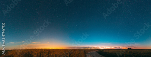 Night Starry Sky With Glowing Stars Above Countryside Landscape. Light Cloudiness Overcast Above Rural Field Meadow And Country Road In Summer. Panorama, Panoramic View.