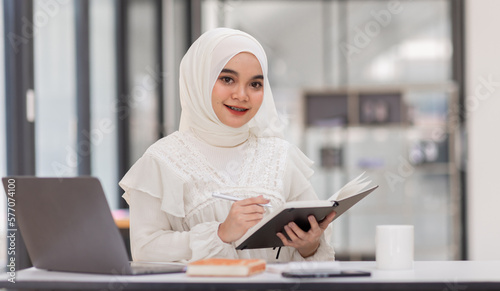 Student muslim woman, Beautiful Asian woman wearing the Islamic headscarf, sitting on a wooden table holding pen writing on notebook and cute smile in workplace with computer.