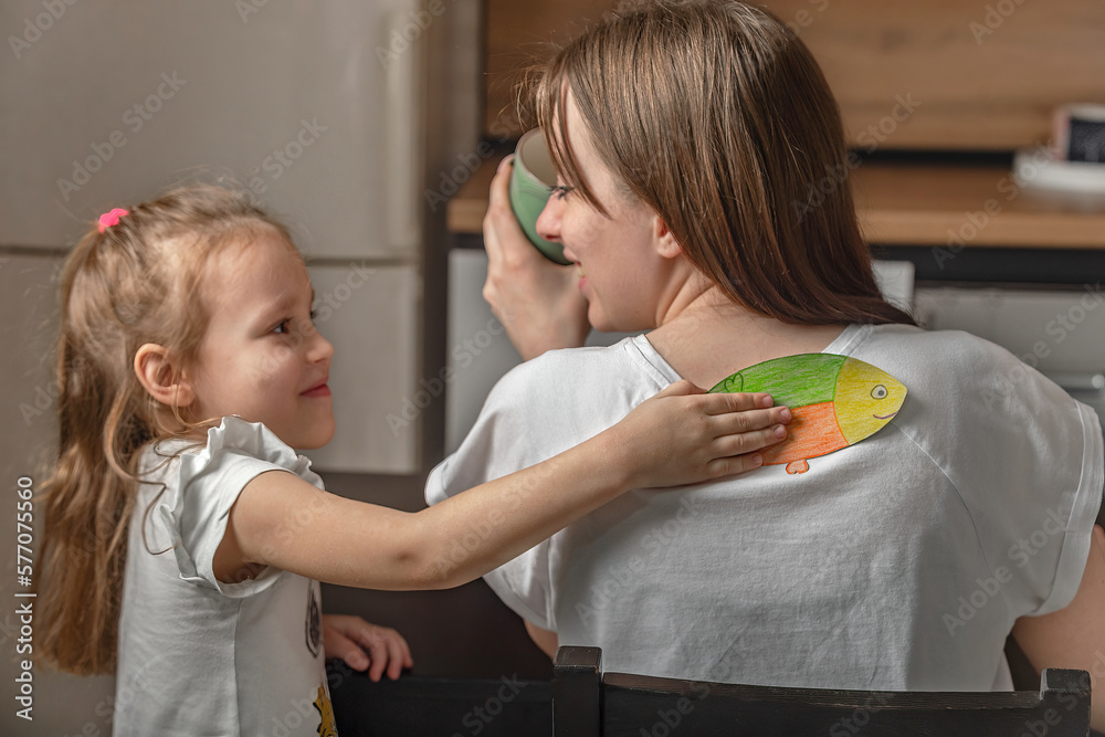 Cheerful little girl glued a piece of paper fish to her mother on her back at home in the kitchen. Funny family jokes, April Fool's Day celebration - obrazy, fototapety, plakaty 