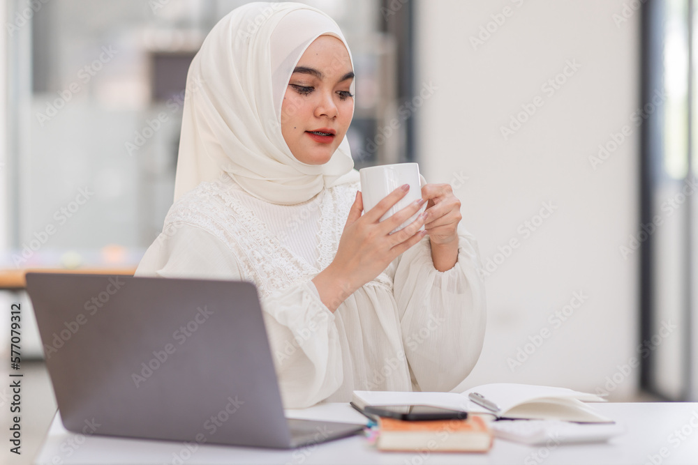 Work With Comfort. Portrait Of Cute Smile Muslim Woman In Hijab Drinking Coffee At Desk And Working On Laptop In Workplace Office. Work With Comfort. Portrait Of Cute Smile Muslim Woman In Hijab Drinking Coffee At Desk And Working On Laptop In Workplace Office.