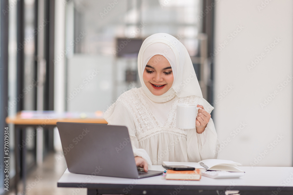 Work With Comfort. Portrait Of Cute Smile Muslim Woman In Hijab Drinking Coffee At Desk And Working On Laptop In Workplace Office.