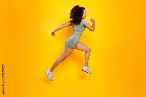 Workout concept. Sports lifestyle. Full length side view photo of a fitness, sporty, motivated mixed race woman in sportswear, training working out, jumping, running, isolated yellow background
