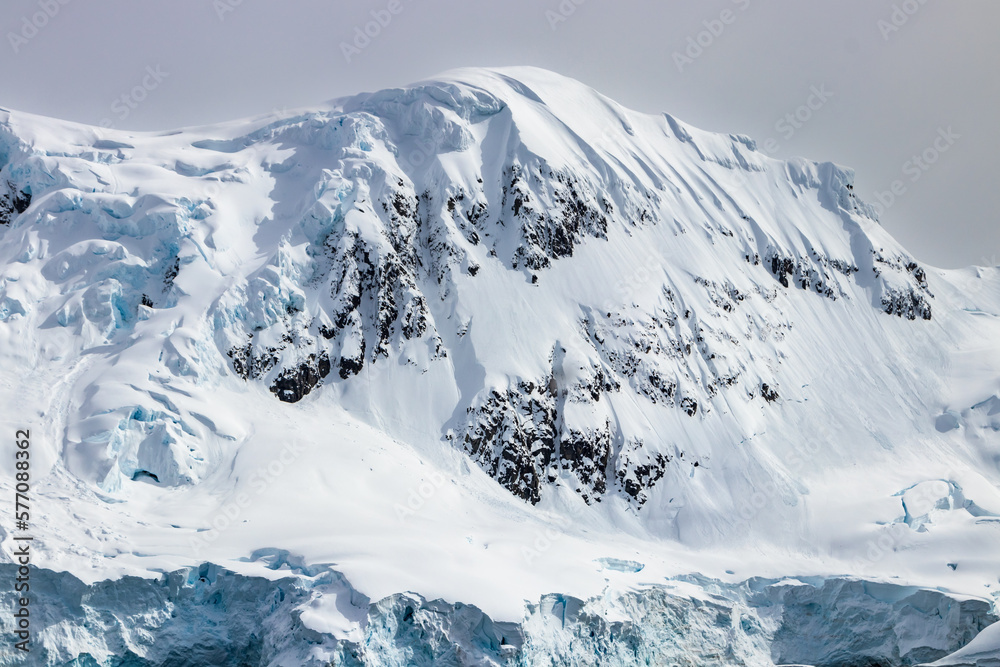 Snow covered mountain peak, Antarctic Peninsula. Rocks exposed on snowy slope. Blue ice visible. Cloudy sky in background. 
