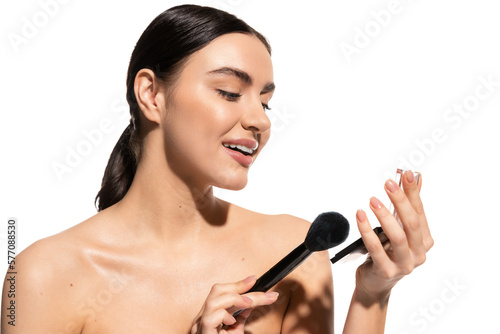 cheerful woman with bare shoulders holding soft cosmetic brush near powder blush isolated on white.