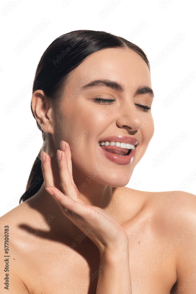 cheerful young woman with natural makeup and bare shoulder touching cheek while sticking out tongue isolated on white.
