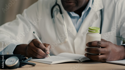 Unknown male doctor african american ethnic man sit at desk with stethoscope write notes prescribe medical insurance checklist writing dosage of medication hold bottle drug antibiotic pills vitamins