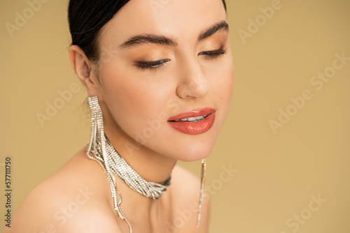 brunette woman in necklace and earrings looking away while posing isolated on beige.