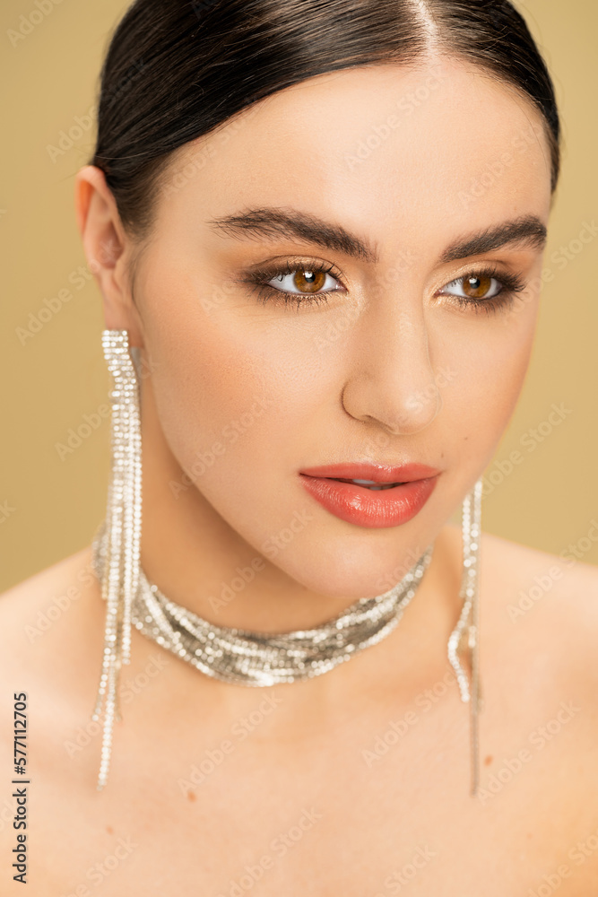 portrait of brunette young woman in necklace and earrings looking away isolated on beige.