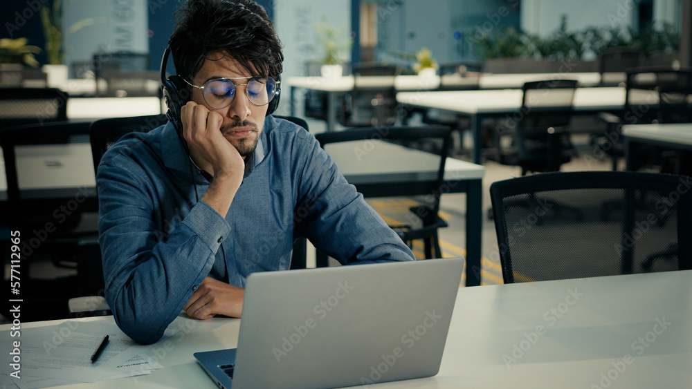 Bored tired arabian indian ethnic man businessman sit at office desk exhausted lazy sad male in headphones listen boring online webinar feel boredom lack of motivation dream about end of working day Bored tired arabian indian ethnic man businessman sit at office desk exhausted lazy sad male in headphones listen boring online webinar feel boredom lack of motivation dream about end of working day