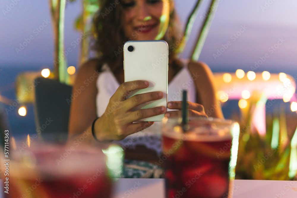 Dark-haired woman taking a picture with her mobile phone of a pair of reddish colored soft drinks on a summer bar terrace with lights and the sea in the background.