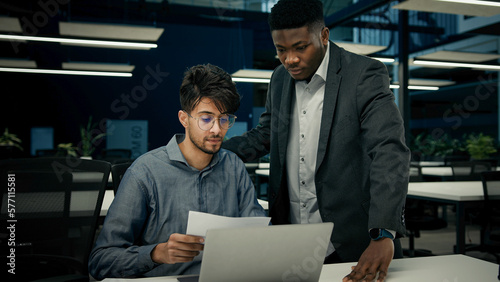 Male African boss businessman mentor teaching indian man trainee new employee discuss online business project on laptop explain details documents to colleague men working together teamwork in office