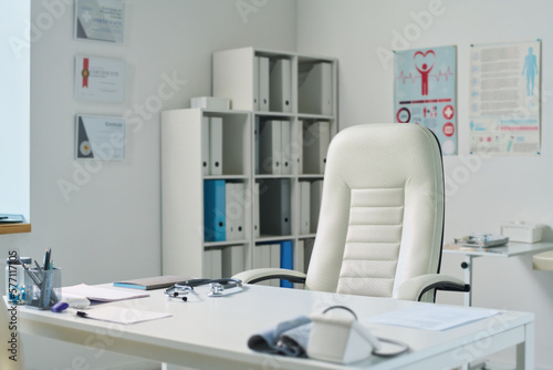 Empty medical office with workplace of clinician consisting of desk and white leather armchair standing against shelves with folders