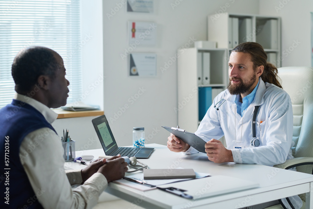© pressmaster - Young doctor in lab coat communicating with African American senior male patient while sitting by workplace in front of him