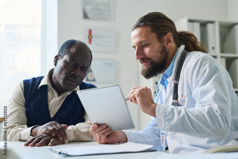 Fototapeta premium Serious mature patient looking at tablet screen and listening to clinician explaining results of medical examination during consultation