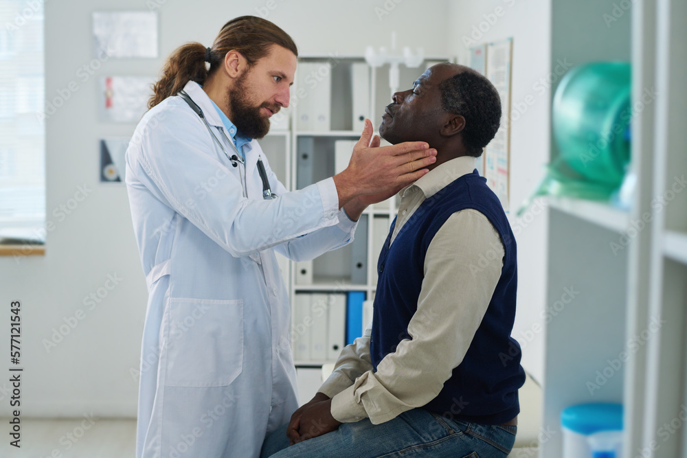 Obraz premium Young endocrinologist checking thyroid of mature African American male patient in casualwear sitting by wall in front of him in hospital