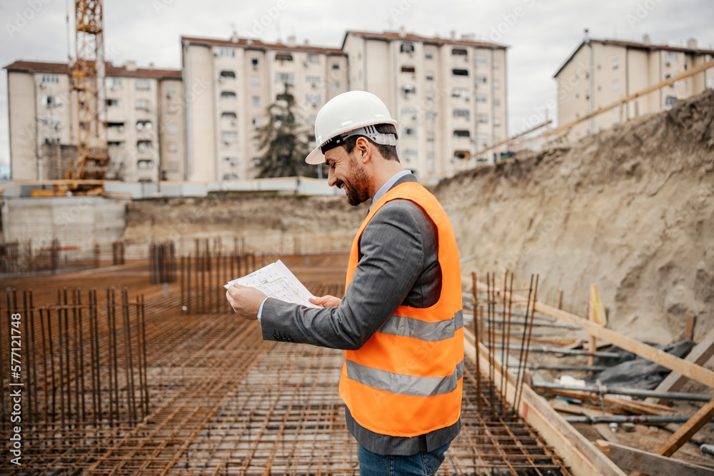 A happy engineer is holding plans and documents and checking on it while standing at construction site.