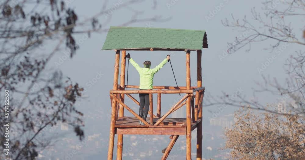 Man standing on a wooden watch tower in the mountains with a view of a city. Raising hands as a gesture of success. Trail runner reaching his destination. Man standing on a wooden watch tower in the mountains with a view of a city. Raising hands as a gesture of success. Trail runner reaching his destination.
