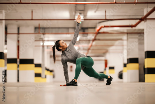 A flexible sportswoman is stretching her legs and backs in underground parking lot.