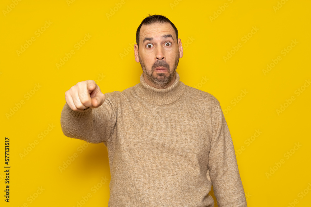 Bearded hispanic man in his 40s wearing a beige turtleneck pointing at something scary, he is very scared and feeling totally terrified. Isolated on yellow background. Bearded hispanic man in his 40s wearing a beige turtleneck pointing at something scary, he is very scared and feeling totally terrified. Isolated on yellow background.
