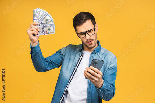 Happy winner! Excited man in casual holding lots of money in dollar currencys and using phone in hands isolated over yellow background.