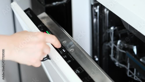 A finger is turning on a built-in dishwasher and selecting eco mode, closing the door.A woman hand is pressing the start button of built-in dishwasher control panel.Modern white kitchen. Close-up	
 photo