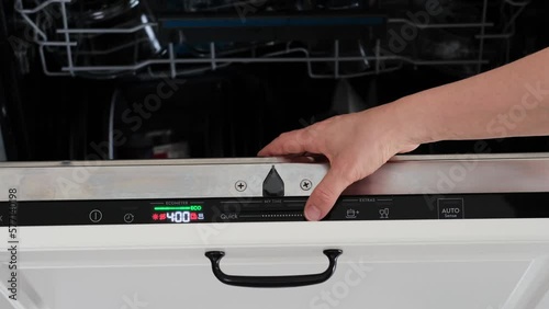 A woman hand is choosing a dishwashing program on a built-in dishwasher. Dishwasher panel. eco mode. A woman hand is pressing the start button of built-in dishwasher control panel.Modern white kitchen