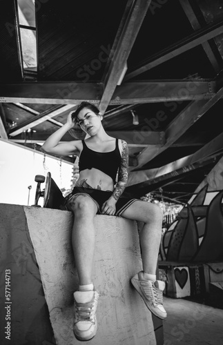 Rebel style: sexy young woman with black top and short, tattoos and skateboard in urban skate park with graffitis (in black and white)