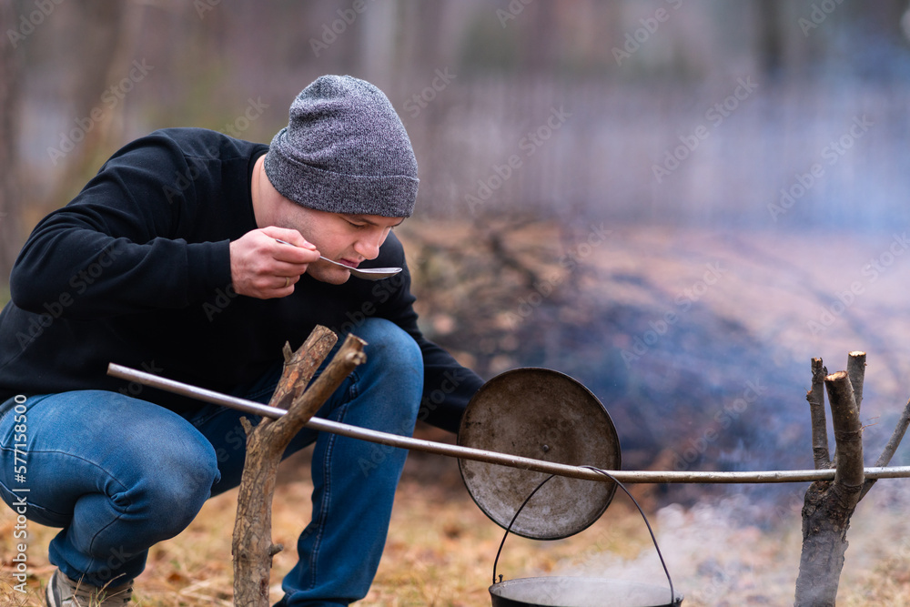 A young guy tastes the broth being cooked over a campfire