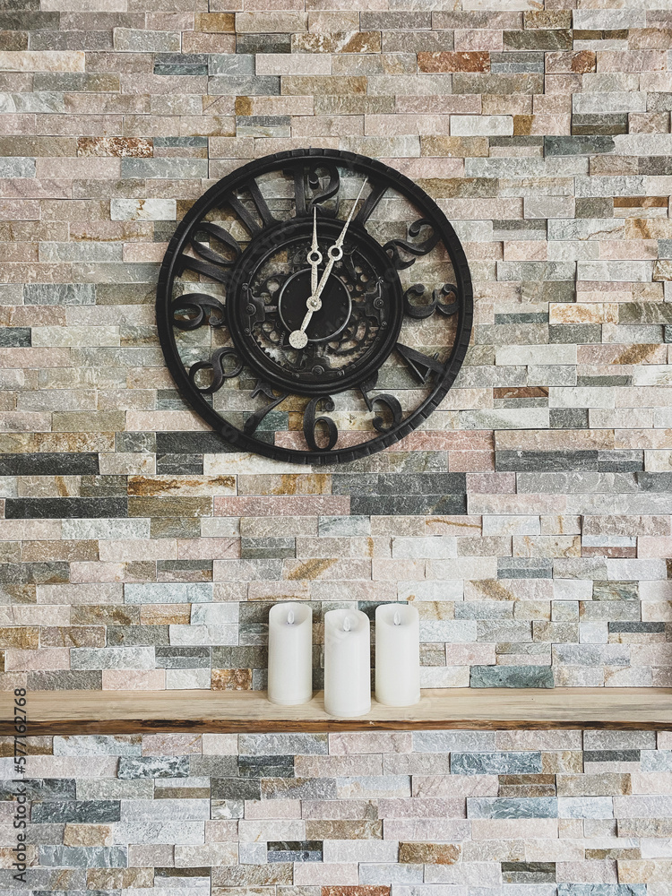 Wrought iron clock on a brick wall above a wooden mantle with three candles