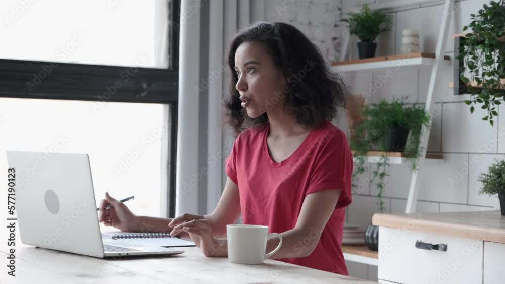 Young African American woman has video call using laptop for freelance work at kitchen. Curly-haired female in red T-shirt explains online waving arms, makes notes in paper notebook at home