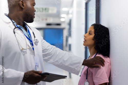 Exhausted diverse female doctor with stethoscope leaning on wall with male doctor with tablet photo