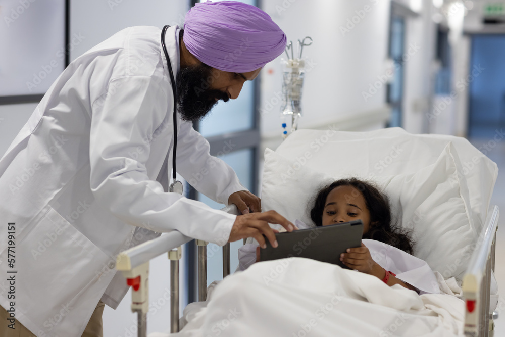 Sikh male doctor in turban with girl using tablet and lying on hospital bed