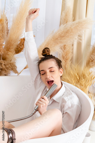 Beautiful teenage girl is sitting in bathtub and singing using shower head, having fun.