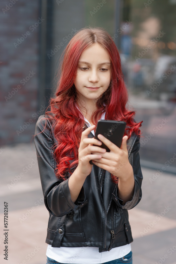Young beautiful girl with red hair in casual clothes communicating by her smartphone, typing, chatting or working online at city street in front of glass window, lifestyle urban outdoor portrait