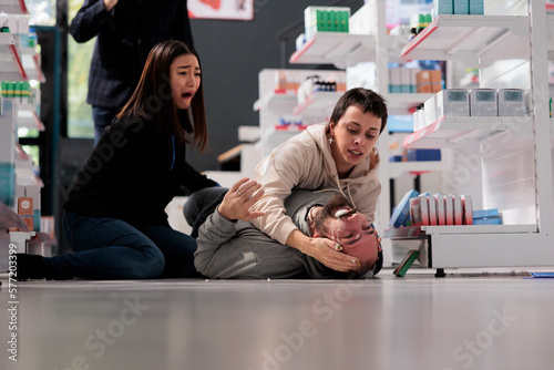 Women crying and screaming for help during person epileptic seizure in medical shop. Young man falling on floor in drugstore, having epilepsy disorder clonic stage symptoms photo