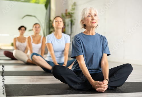 Calm European old female gym visitor practicing seated Bound Angle Pose or Butterfly Pose Baddha Konasana during group yoga training