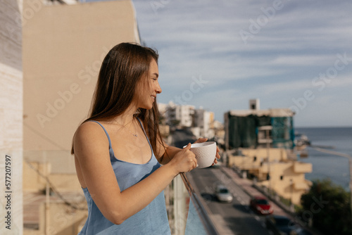Attractive cute stylish European woman with dark hair wearing blue suit is drinking morning coffee on balcony in sunlight. 