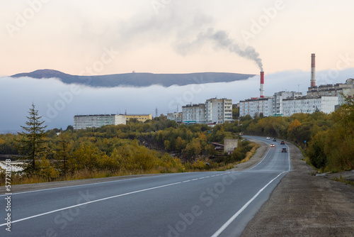 Fototapeta Naklejka Na Ścianę i Meble -  View of the paved road, residential city block and pipes of the combined heat and power plant. Fog and mountains in the distance. City of Magadan, Magadan region, Far East of Russia. Autumn season.