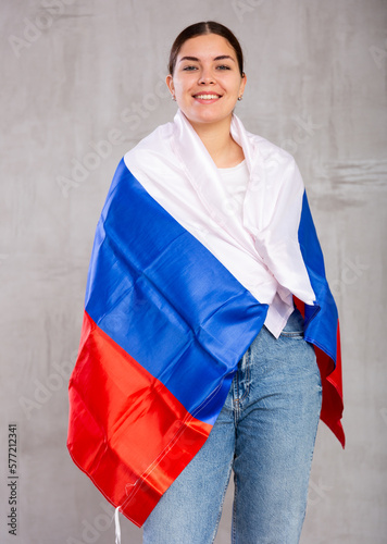 Smiling young woman in casual clothes wrapping herself in Russian flag against gray wall background