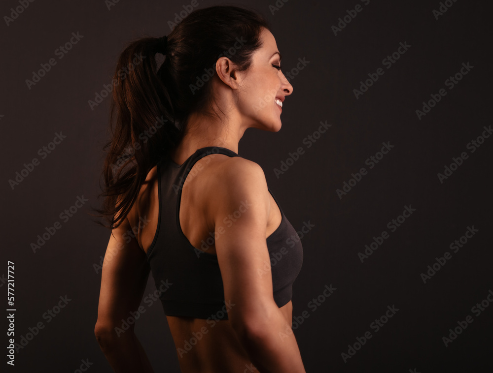 Naklejka premium Smiling happy female sporty muscular with ponytail with stretching workout of shoulders, blades and arms in sport bra, standing on dark grey background with empty copy space. Back view.
