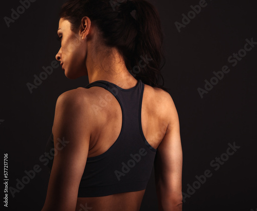 Serious female sporty muscular with ponytail doing stretching workout the shoulders, blades and arms in sport bra, standing on dark grey background with empty copy space. Back view. © nastia1983