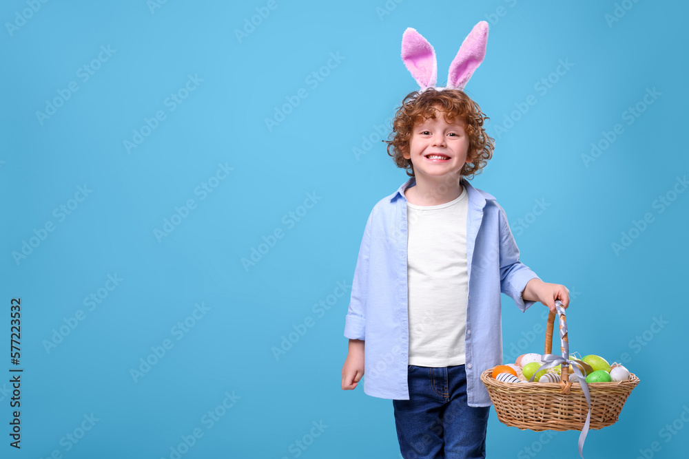 Happy boy in cute bunny ears headband holding wicker basket with Easter eggs on light blue background. Space for text Happy boy in cute bunny ears headband holding wicker basket with Easter eggs on light blue background. Space for text