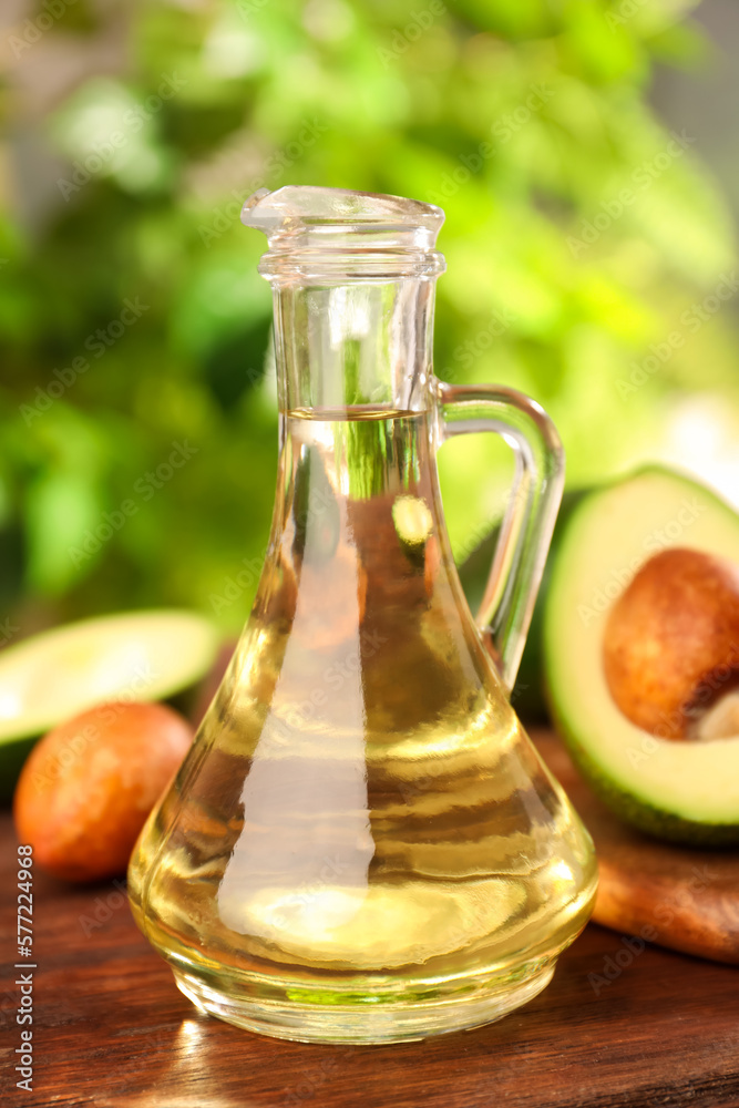 Glass jug of cooking oil and fresh avocados on wooden table against blurred green background Glass jug of cooking oil and fresh avocados on wooden table against blurred green background