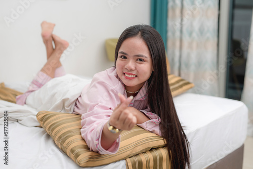 A cute young asian woman in pink pajamas laying happily on the edge of the bed and making a heart finger sign.